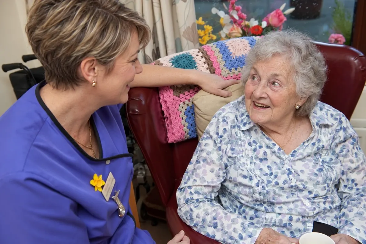 A carer crouched beside a resident in her armchair, the two of them smiling at each other