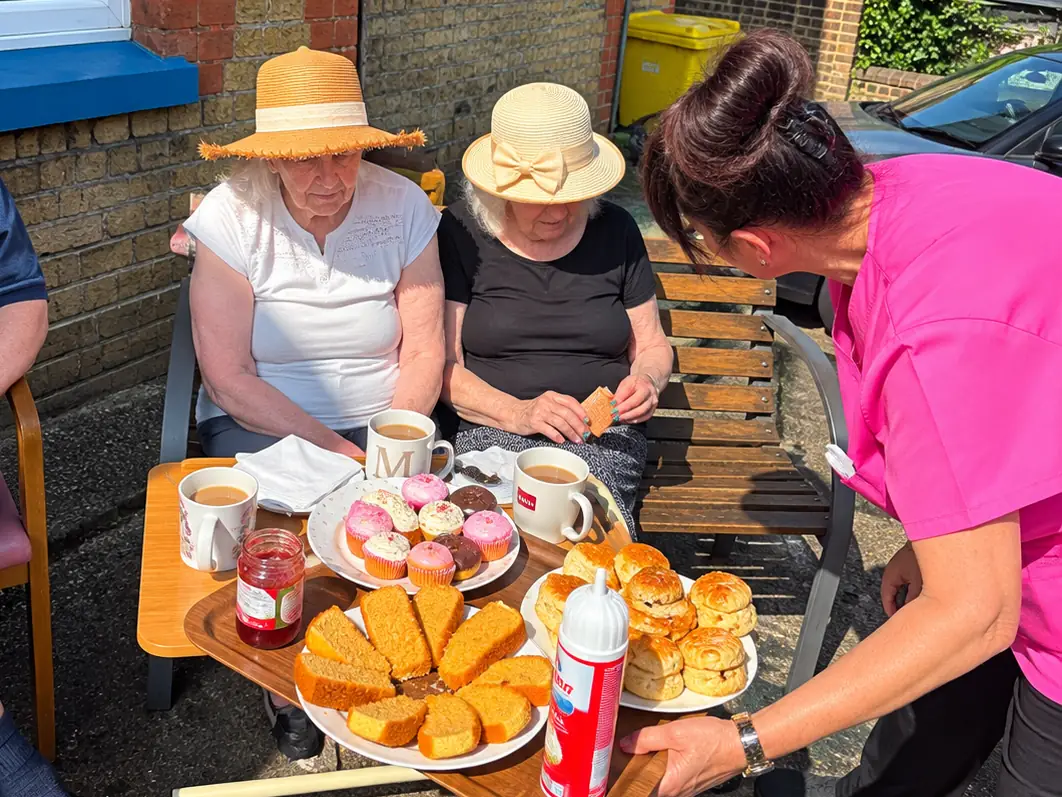 Two residents in straw sun hats on a bench beside the house as a carer brings a tray of tea, cakes, scones, jam and cream