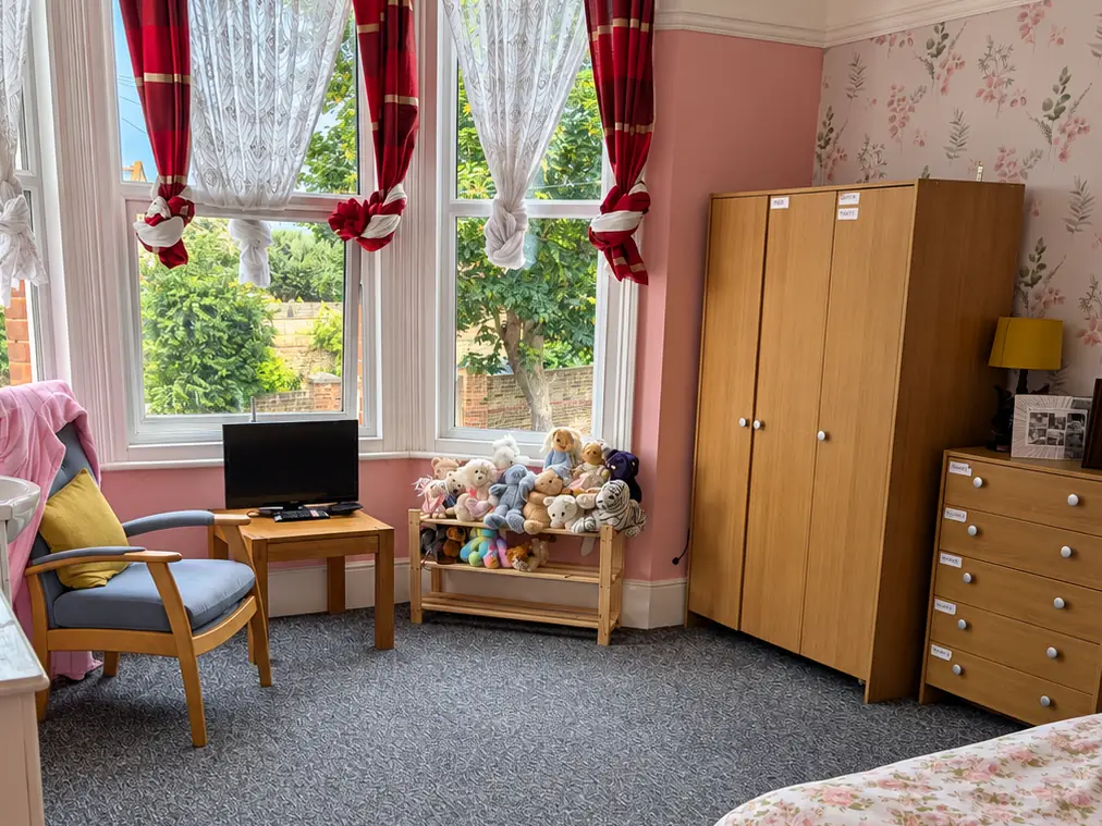 A front bedroom with a bay window and red curtains, a wooden bed with floral bedding, a wardrobe, an armchair, a television and a shelf of soft toys