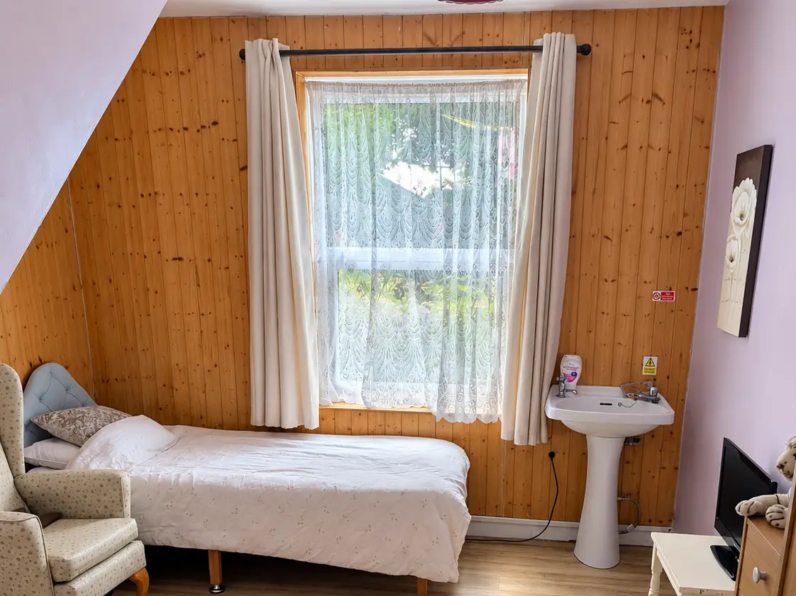 An upstairs bedroom with a sloped ceiling and a pine-clad wall, a made-up single bed, an armchair and a window onto trees